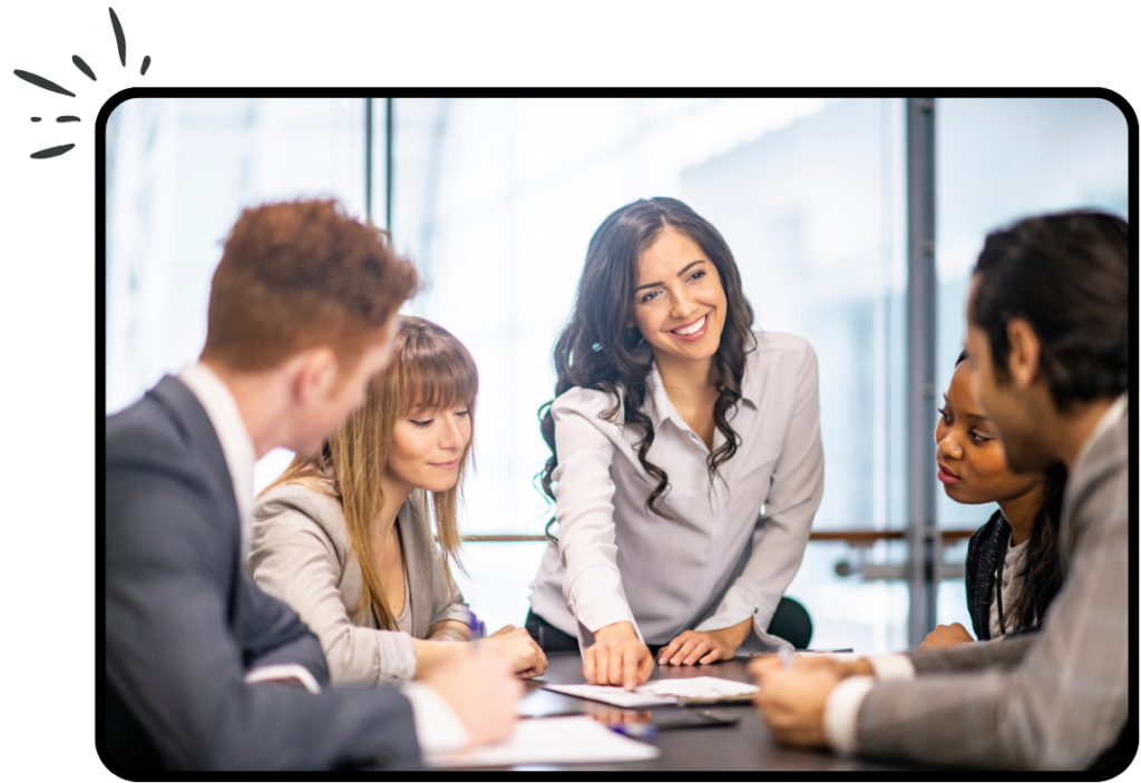 A woman pointing to a piece of paper in the middle of the table with work colleagues sat around.